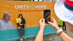 Catholic faithful stand in front of a poster bearing the motto of the papal visit