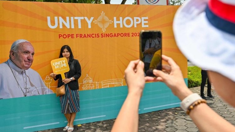 Catholic faithful stand in front of a poster bearing the motto of the papal visit