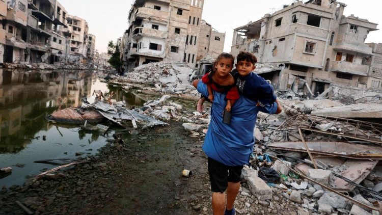 Um homem palestino segura seus filhos enquanto caminha próximo a edifícios destruídos por um ataque israelense, em Khan Younis